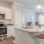 Fully equipped kitchen at Sanctuary at Powell Place in Pittsboro, NC, featuring a large island, pendant lighting, built-in microwave, stove, white cabinetry, wood-like plank flooring, and open living area with ceiling fan.